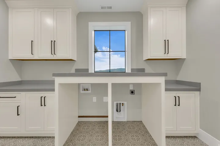 Laundry room featuring a large window, patterned floor tile, extensive cabinetry with dark hardware, and a built-in counter for appliances
