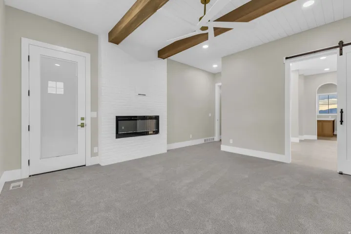 Spacious room featuring vaulted ceilings with exposed wood beams, a contemporary linear fireplace set into a white brick wall, and plush gray carpeting