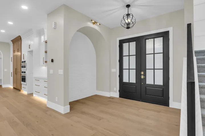 Double black entry doors with frosted glass panels, an arched alcove with white brick-patterned wall, light wood-finish flooring, and a contemporary chandelier