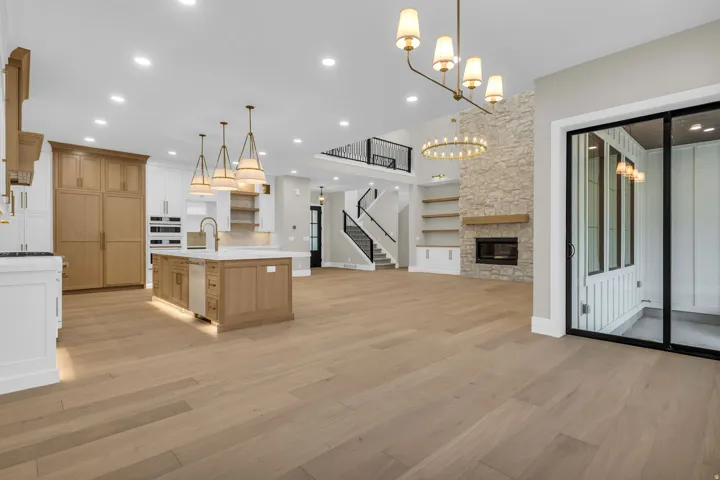 Open-concept living space with light wood-finish flooring, a stone-clad fireplace, a kitchen island with integrated sink, a two-story ceiling, and a sliding glass door