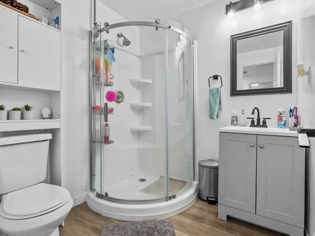 Bathroom featuring a curved glass shower enclosure, a single vanity with a light gray cabinet and white countertop, wood-finish flooring, and white built-in storage cabinetry