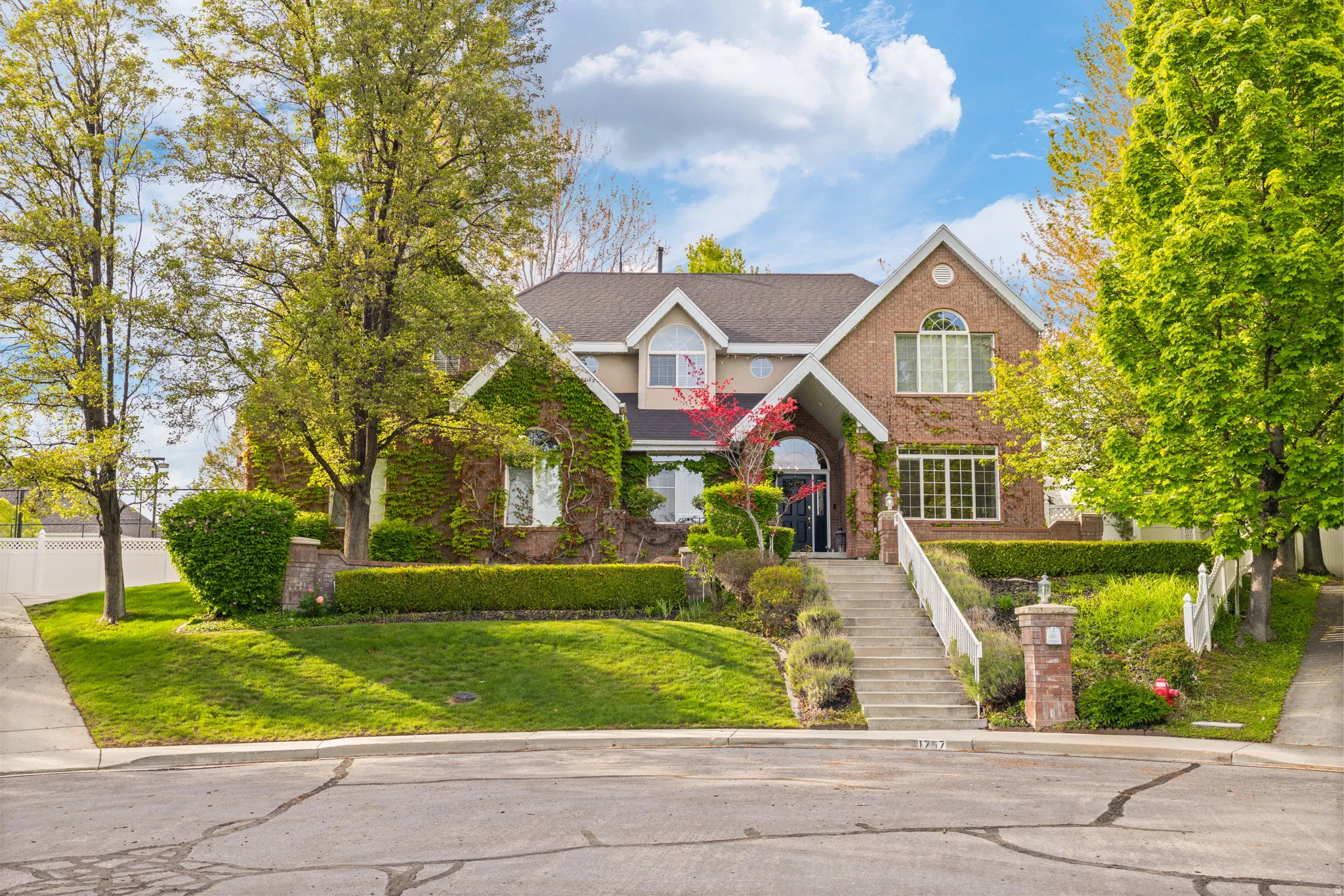 Brick exterior with ivy accents, arched entryway, multi-paned windows, and a tiered landscaped front yard featuring mature trees and a concrete staircase with white railings