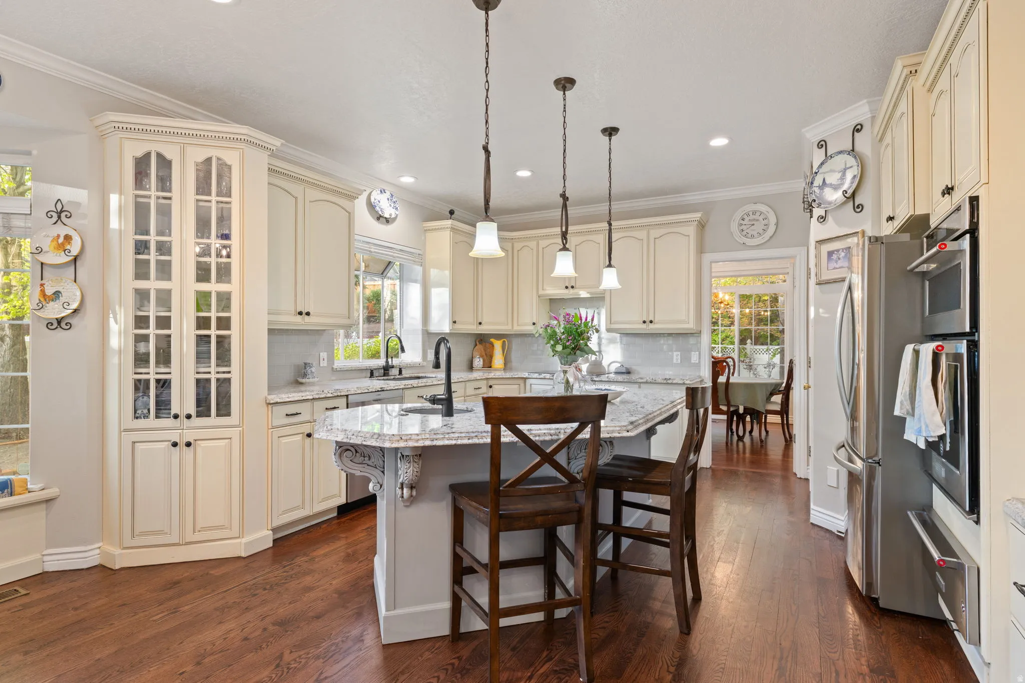 Bright kitchen featuring wood flooring, cream-colored cabinetry with display glass, light granite countertops, and stainless steel appliances including a built-in double oven