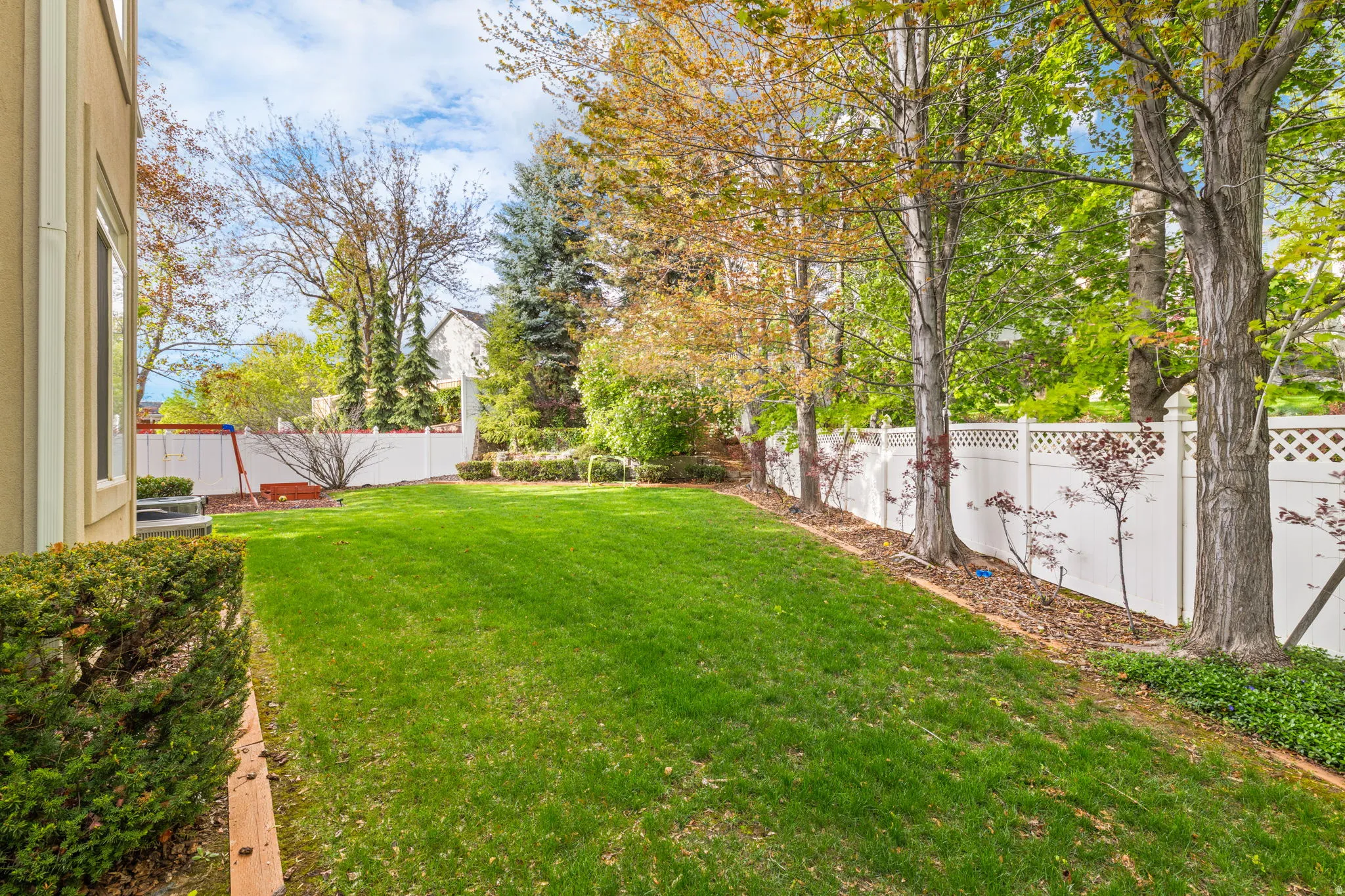 Expansive rear yard featuring a manicured lawn, established perimeter trees, and a white lattice-top privacy fence