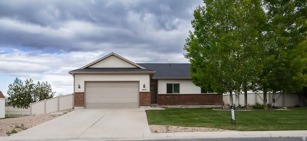 Single-story residence featuring a two-car garage, brick and siding exterior, a front lawn with mature trees, a concrete driveway, and a privacy fence