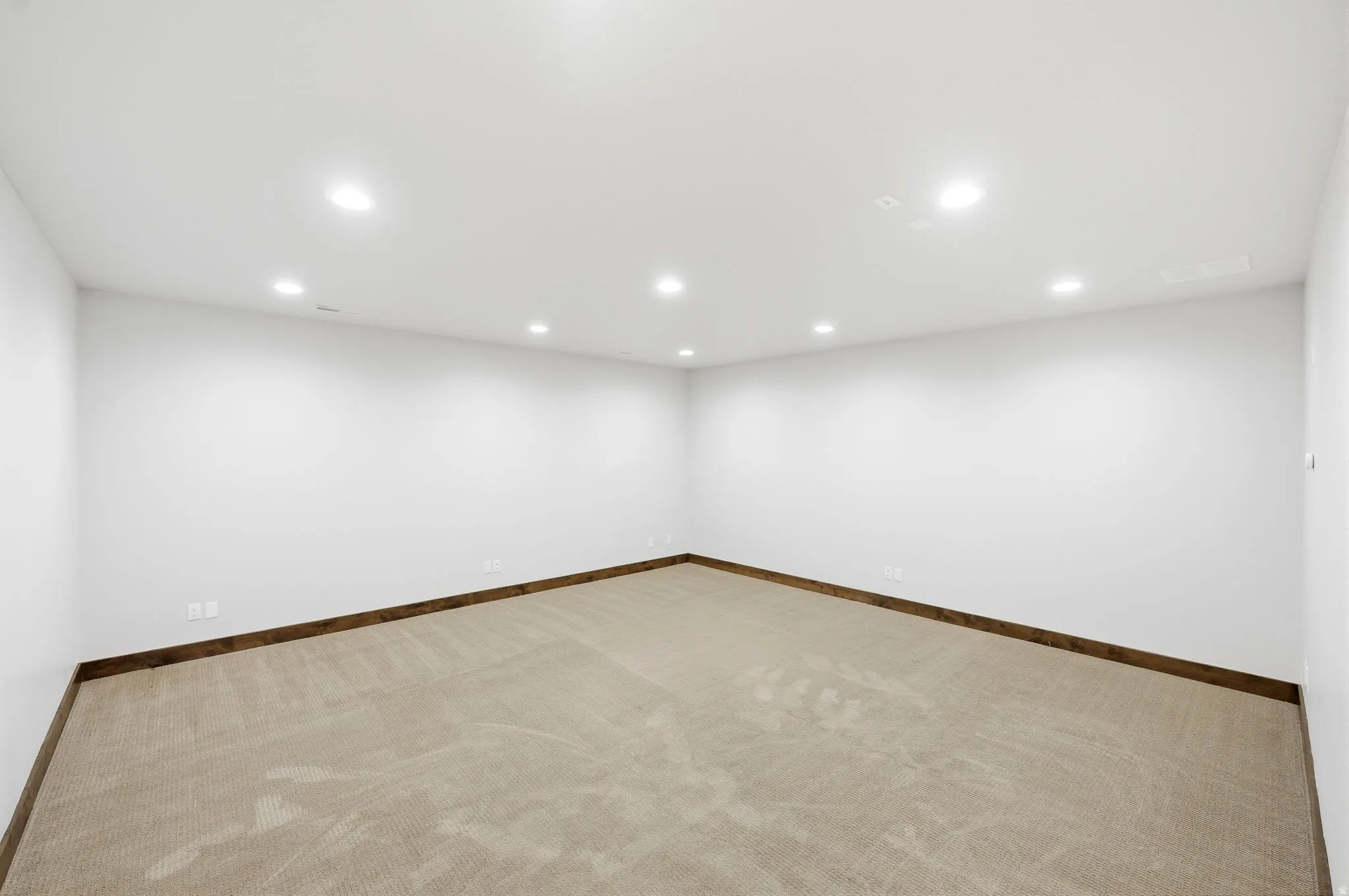 Spacious room with white walls, recessed ceiling lighting, light-toned carpet flooring, and dark wood-finish baseboards