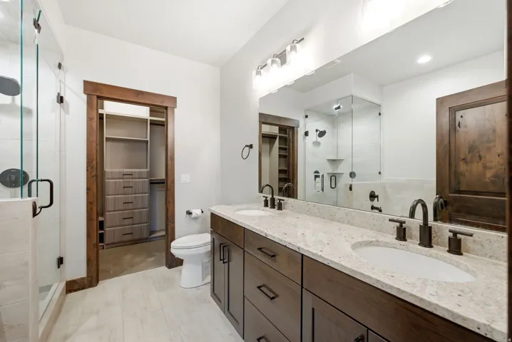 Dual vanity with granite countertops and undermount sinks, matte black fixtures, custom wood cabinetry, glass-enclosed shower, and wood-finish flooring