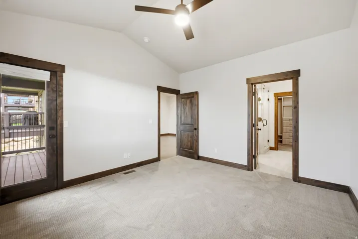 Vaulted ceiling room featuring carpet flooring, dark wood trim, a ceiling fan, and a glass-paneled door opening to an outdoor deck