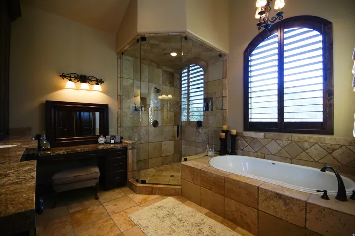 Tiled bathroom featuring a built-in bathtub, a walk-in shower with glass enclosure, a vanity with stone countertop, and arched wood-frame windows with plantation shutters