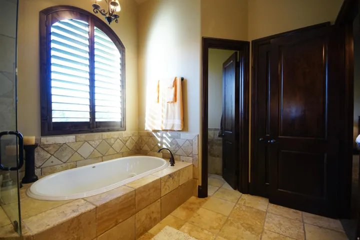 Soaking tub with a tiled surround, arched window featuring wooden plantation shutters, and a frameless glass shower enclosure