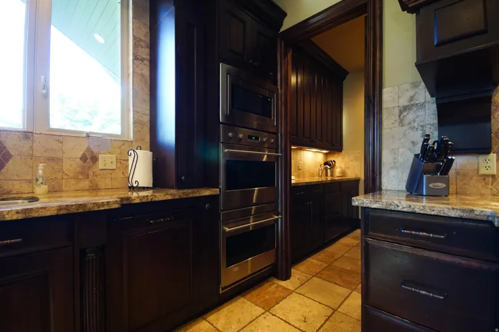 Kitchen featuring dark wood cabinetry, a built-in stainless steel microwave and oven, a neutral-toned tile backsplash with accent tiles, light-toned countertops, and tile flooring