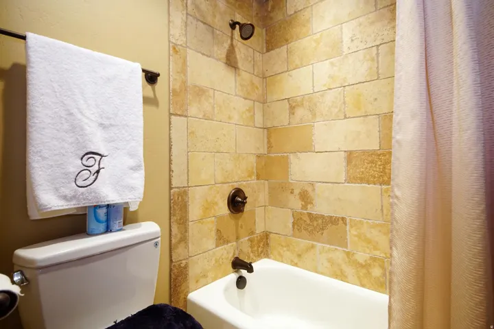 Bathtub with a shower fixture, stone tile surround, and bronze hardware