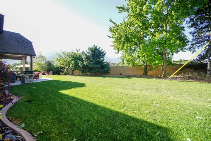 Expansive backyard featuring a manicured lawn, established landscaping, mountain scenery, concrete patio, and a covered outdoor living area