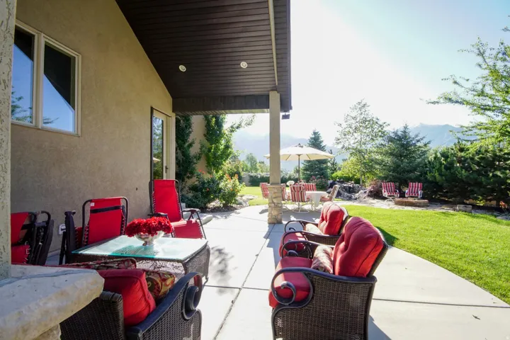 Covered concrete patio extending to a manicured lawn, featuring a stone-clad pillar and a dark wood-finish ceiling with recessed lighting
