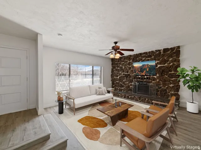 Living area featuring a stone accent wall with a fireplace, a large window, a ceiling fan, and wood-finish flooring