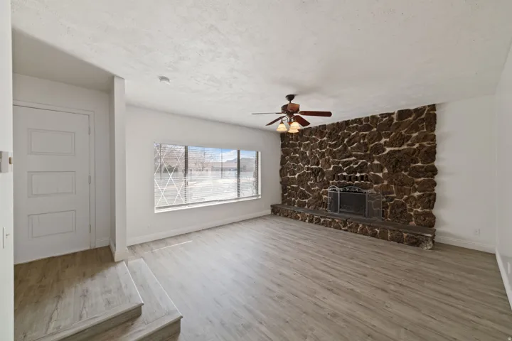 Spacious living area featuring a prominent stone fireplace, wood-finish flooring, a ceiling fan with integrated lighting, a large window, and a paneled interior door
