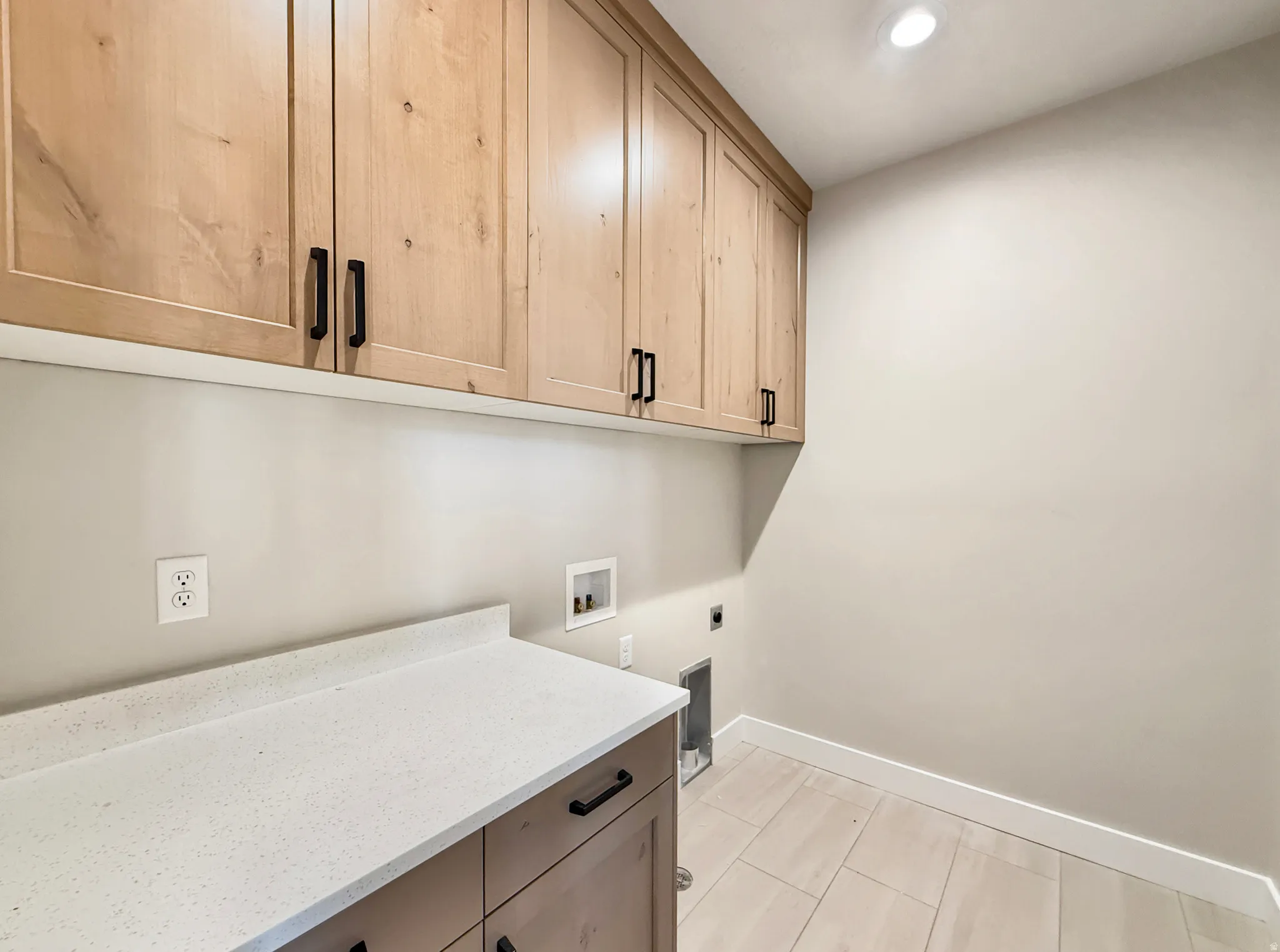 Laundry area featuring light wood-finish cabinetry with matte black hardware, a light speckled countertop, and light-toned tile flooring