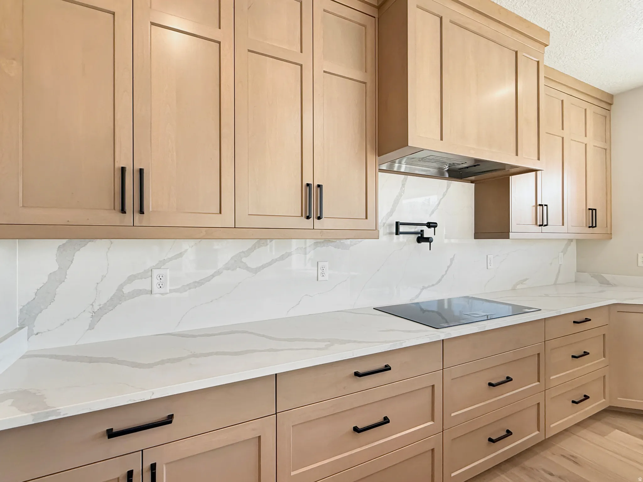 Kitchen featuring extensive light wood-finish cabinetry with black hardware, white countertops and backsplash with grey veining, a built-in cooktop, and an integrated range hood