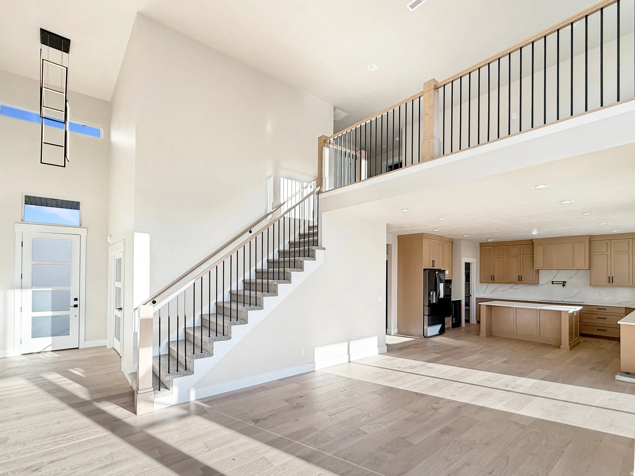 Open-concept living space featuring high ceilings, wood-finish flooring, a contemporary black metal chandelier, and a staircase with light wood handrails and black balusters