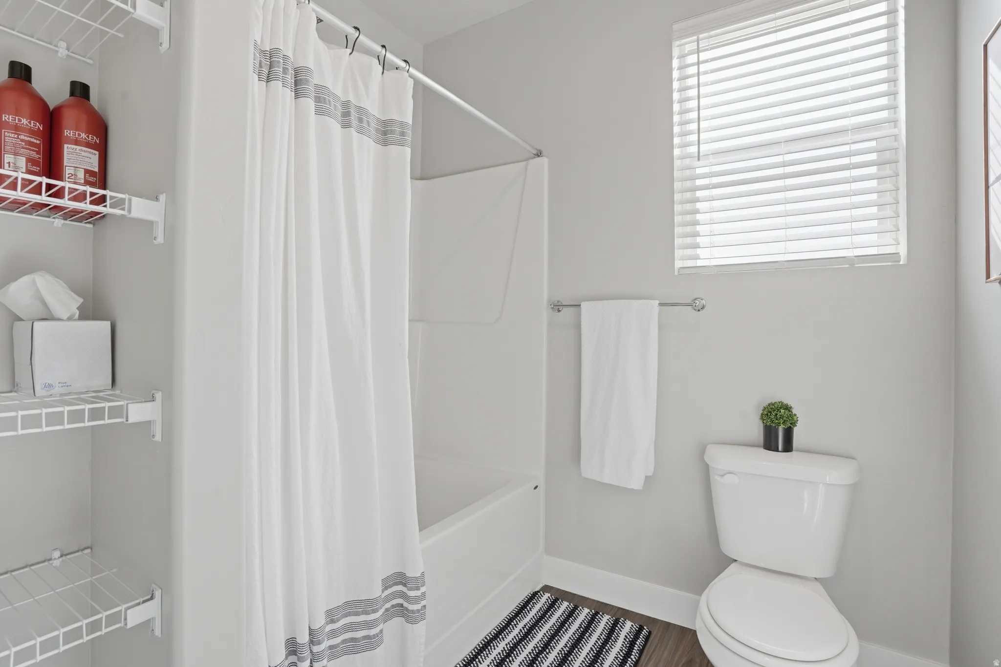 Full bathroom featuring a white tub-shower combination, a toilet, a window with horizontal blinds, and a built-in wire shelving unit