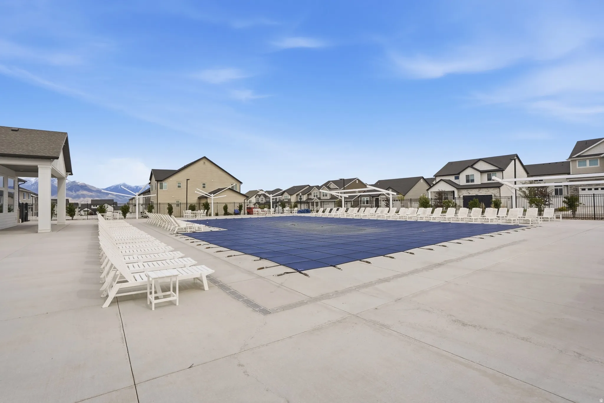 Community pool with extensive surrounding concrete decking, multiple white lounge chairs, and a covered pavilion