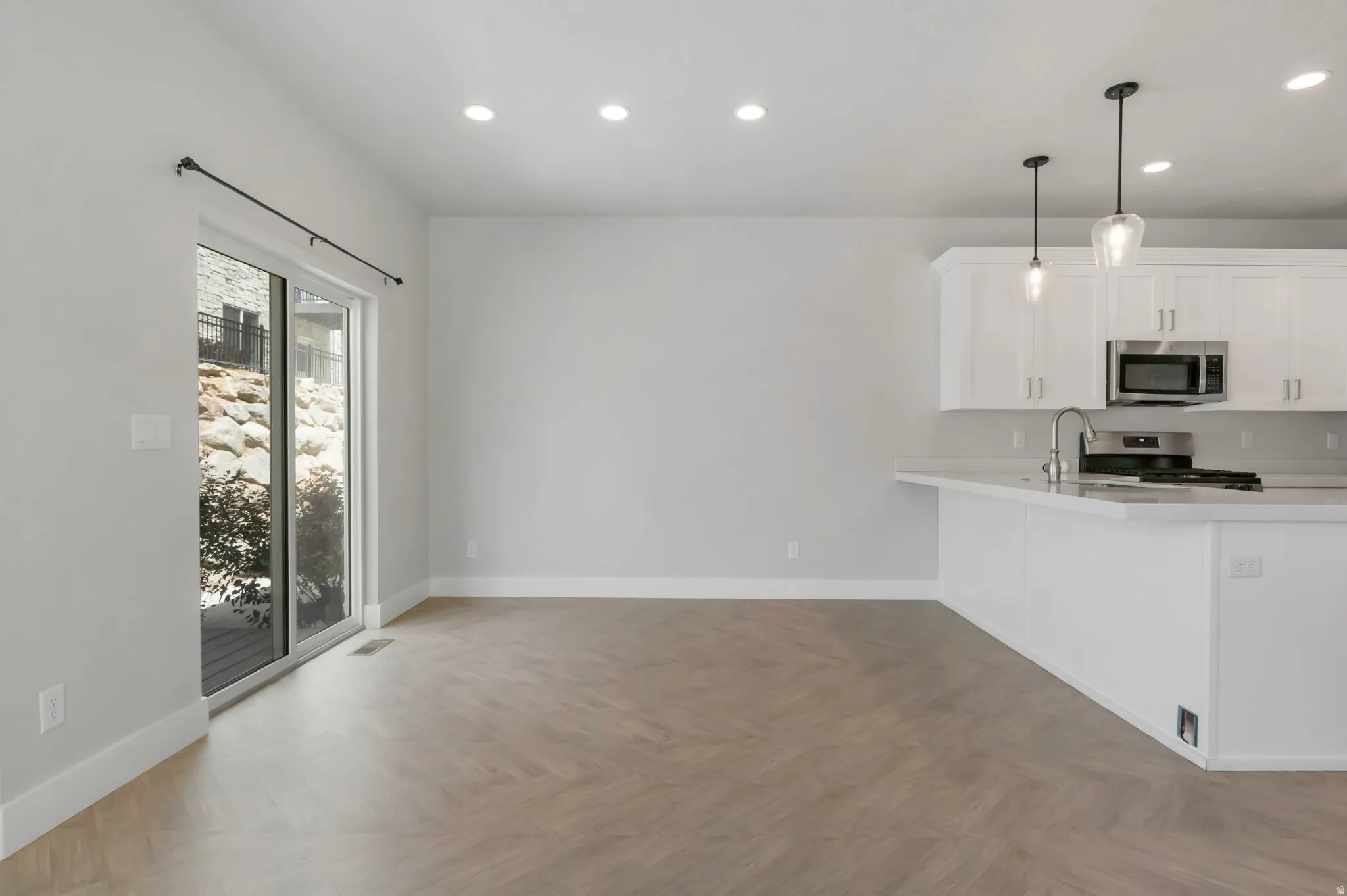 Open-concept interior featuring light wood-finish herringbone flooring, a white kitchen island with a stainless steel sink and faucet, white cabinetry, recessed lighting, and a sliding glass door