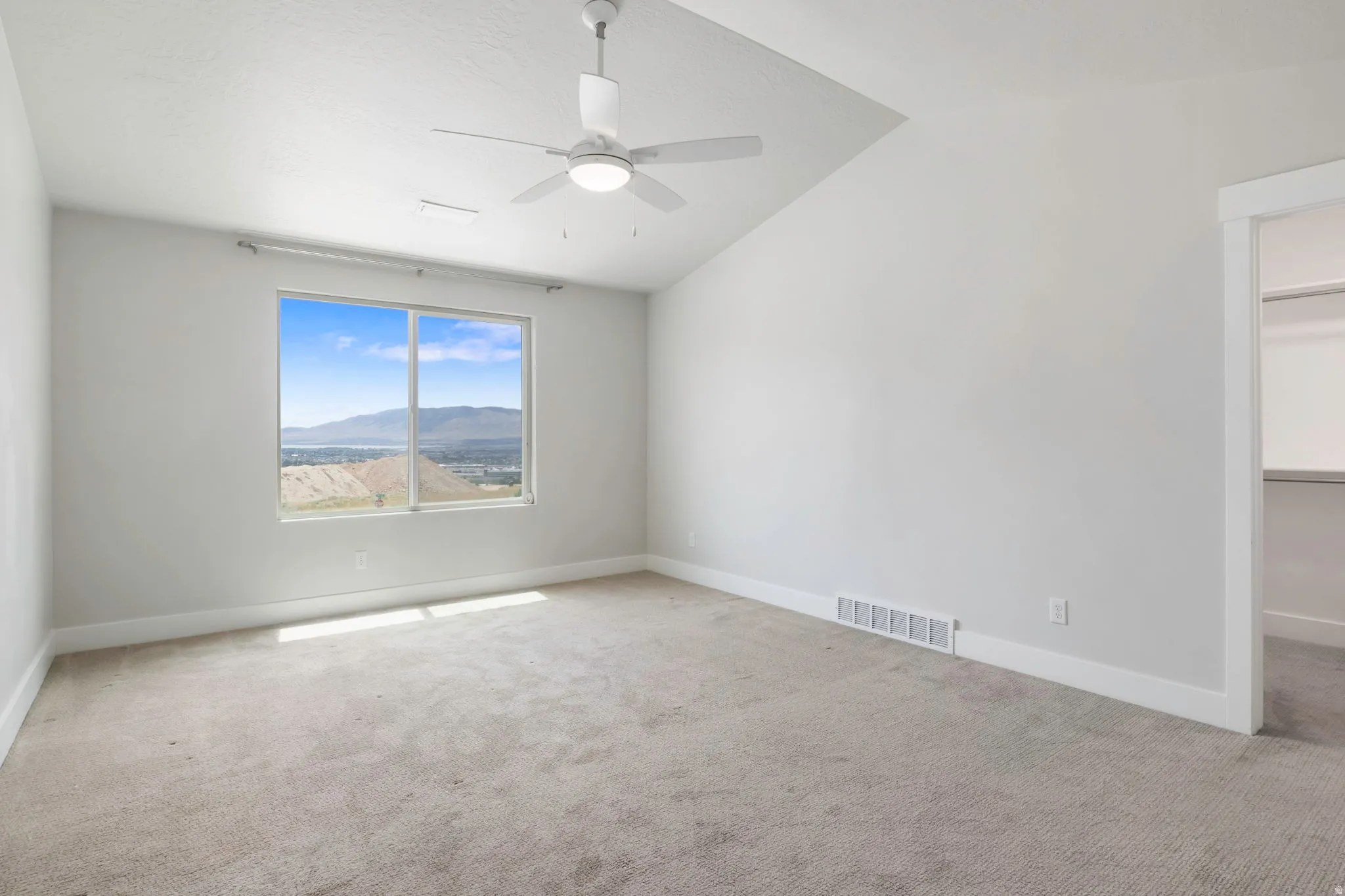 Spacious room featuring a large window with panoramic mountain and water scenery, sloped ceiling, ceiling fan with integrated lighting, and neutral carpet flooring