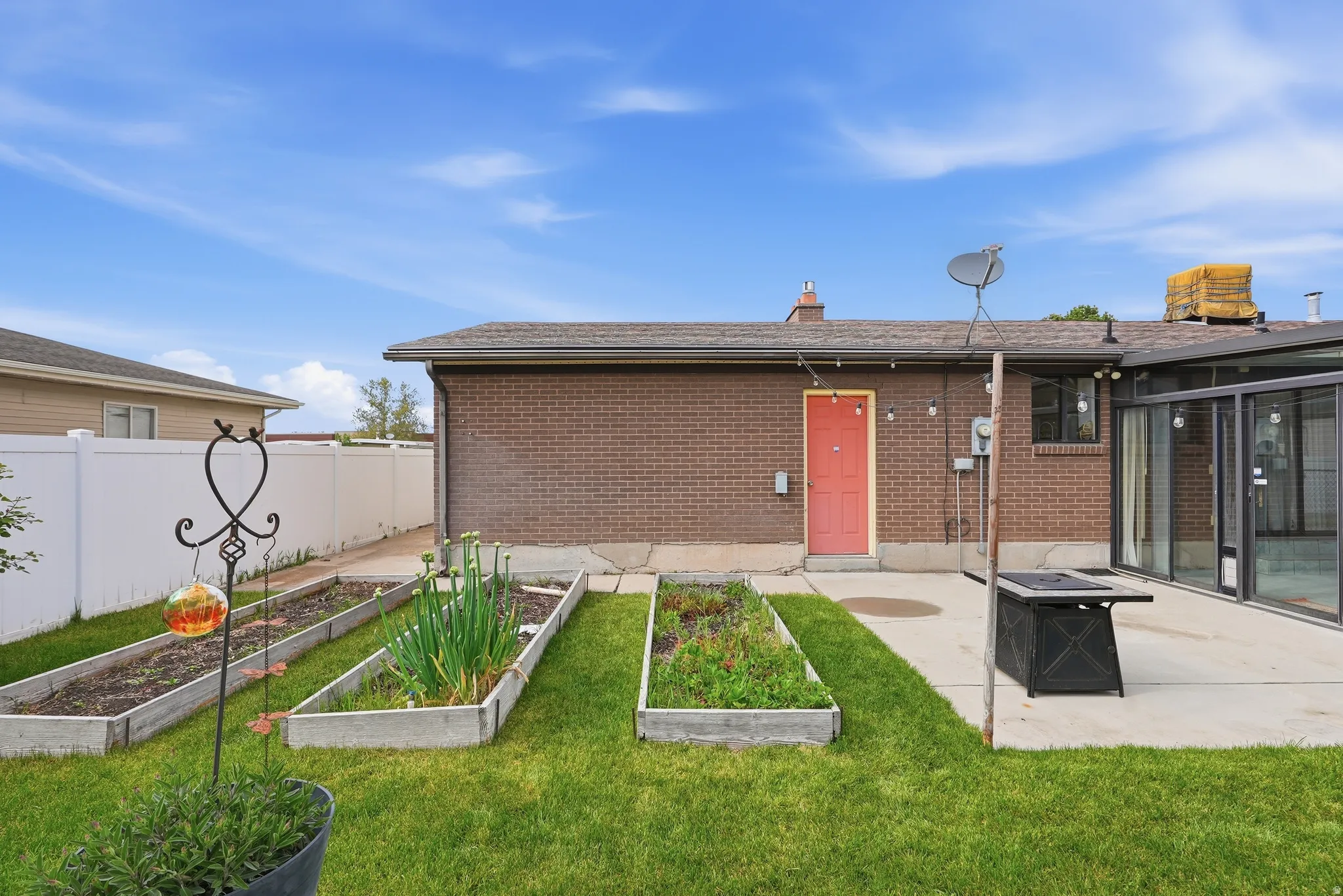 Manicured lawn area featuring multiple raised garden beds, a concrete patio, and a brick exterior with a red entry door
