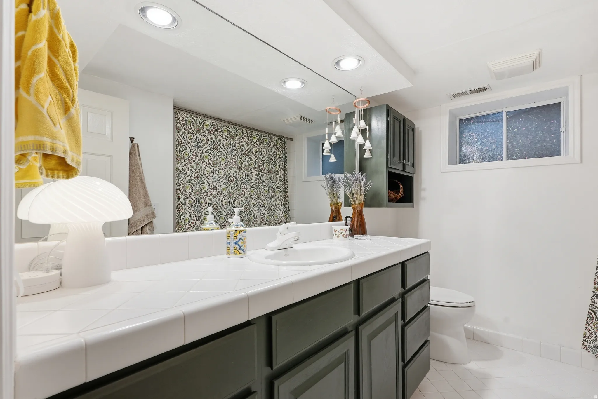 Bathroom featuring a single basin vanity with white tiled countertop, dark painted cabinetry, and a full-width mirror
