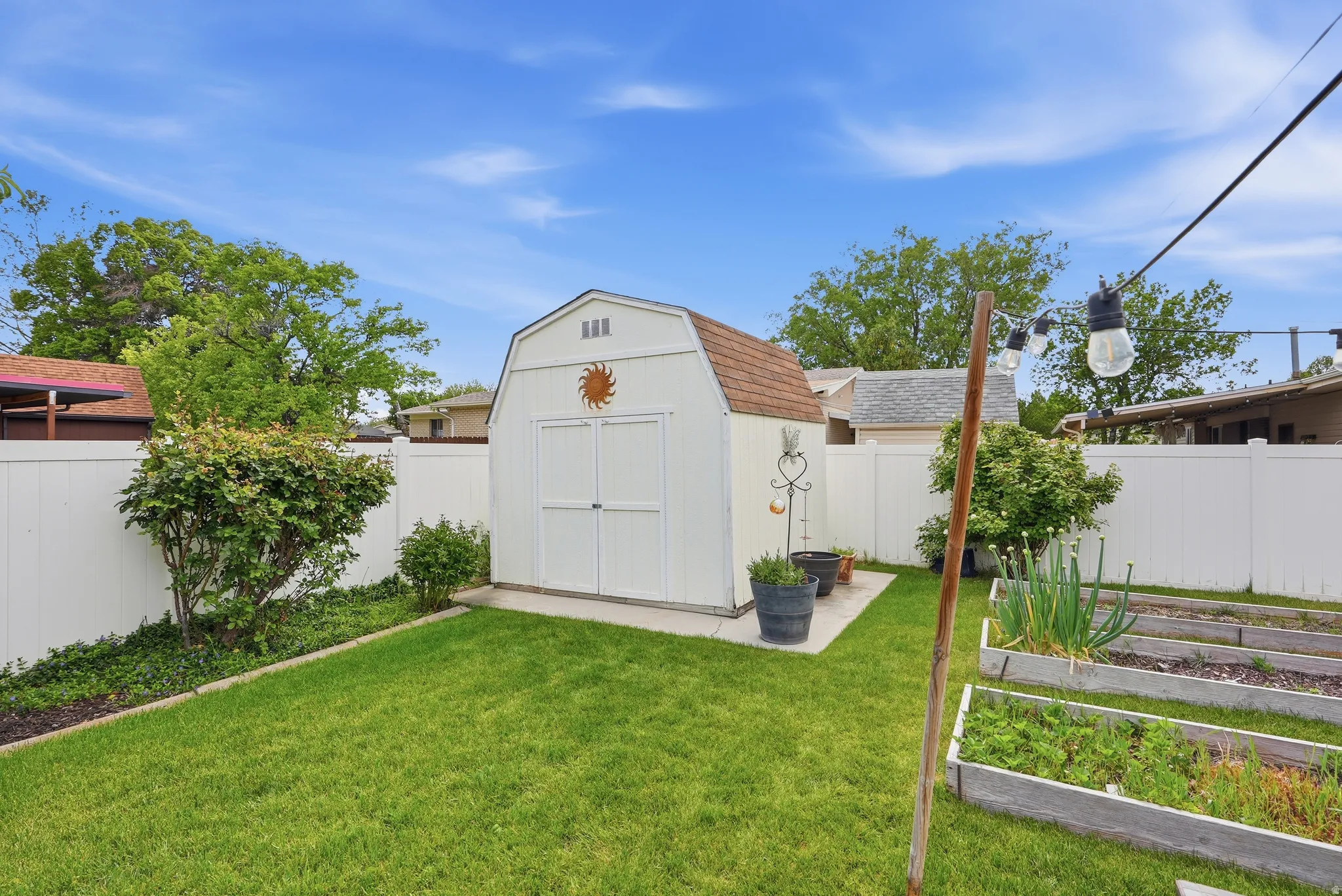 Expansive backyard featuring a manicured lawn, a detached storage shed with a gambrel roof, white privacy fencing, and raised garden beds