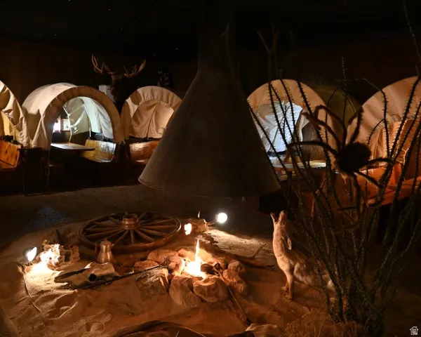 Indoor immersive space featuring multiple covered wagon structures, a central simulated campfire with surrounding rocks, a wagon wheel element, and a large decorative hood