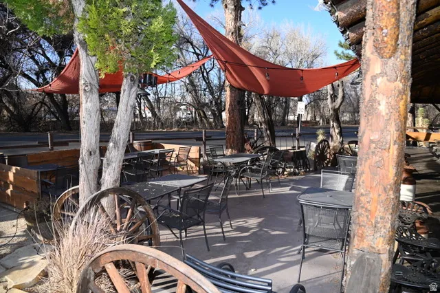 Outdoor patio area featuring multiple seating arrangements, two overhead fabric shade structures, a prominent log column, and decorative wagon wheels