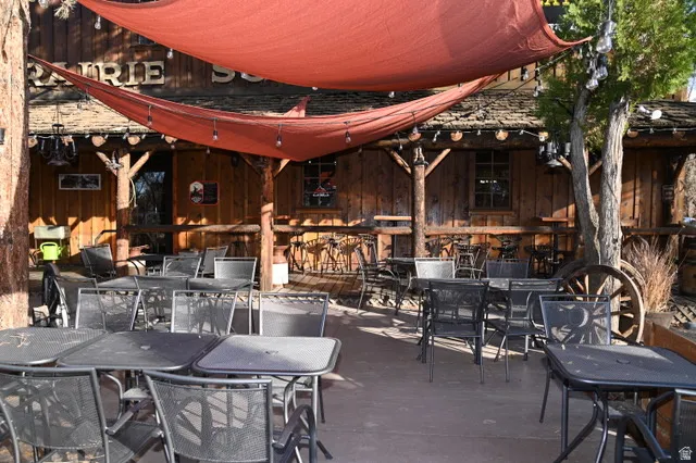 Outdoor dining area featuring multiple metal tables and chairs, overhead red sun shades with string lighting, and a rustic wood-sided building with exposed beam details