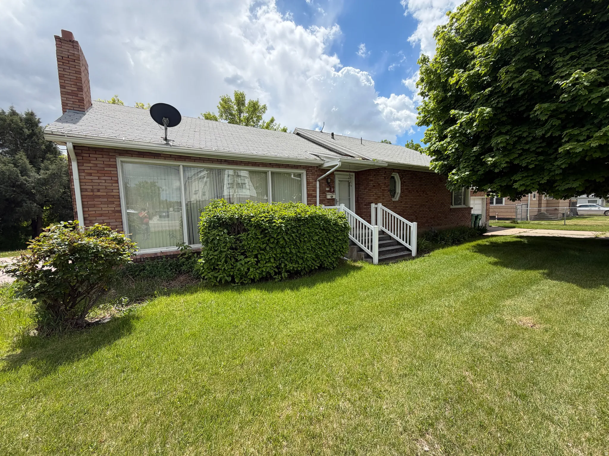 Brick exterior residence featuring a prominent brick chimney, white-framed picture windows, a covered entry with white railings, and mature landscaping
