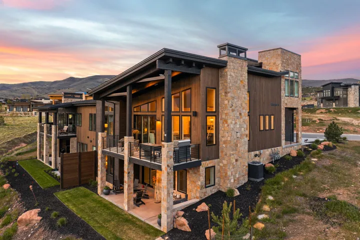 Contemporary exterior featuring stacked stone and dark wood siding, multiple balconies with black railings, expansive windows, a covered patio, and structured landscaping with rock accents