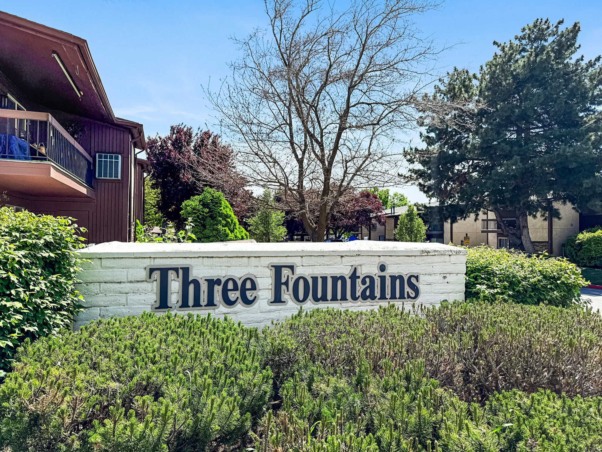 Brick sign with "Three Fountains" lettering, surrounded by varied green foliage