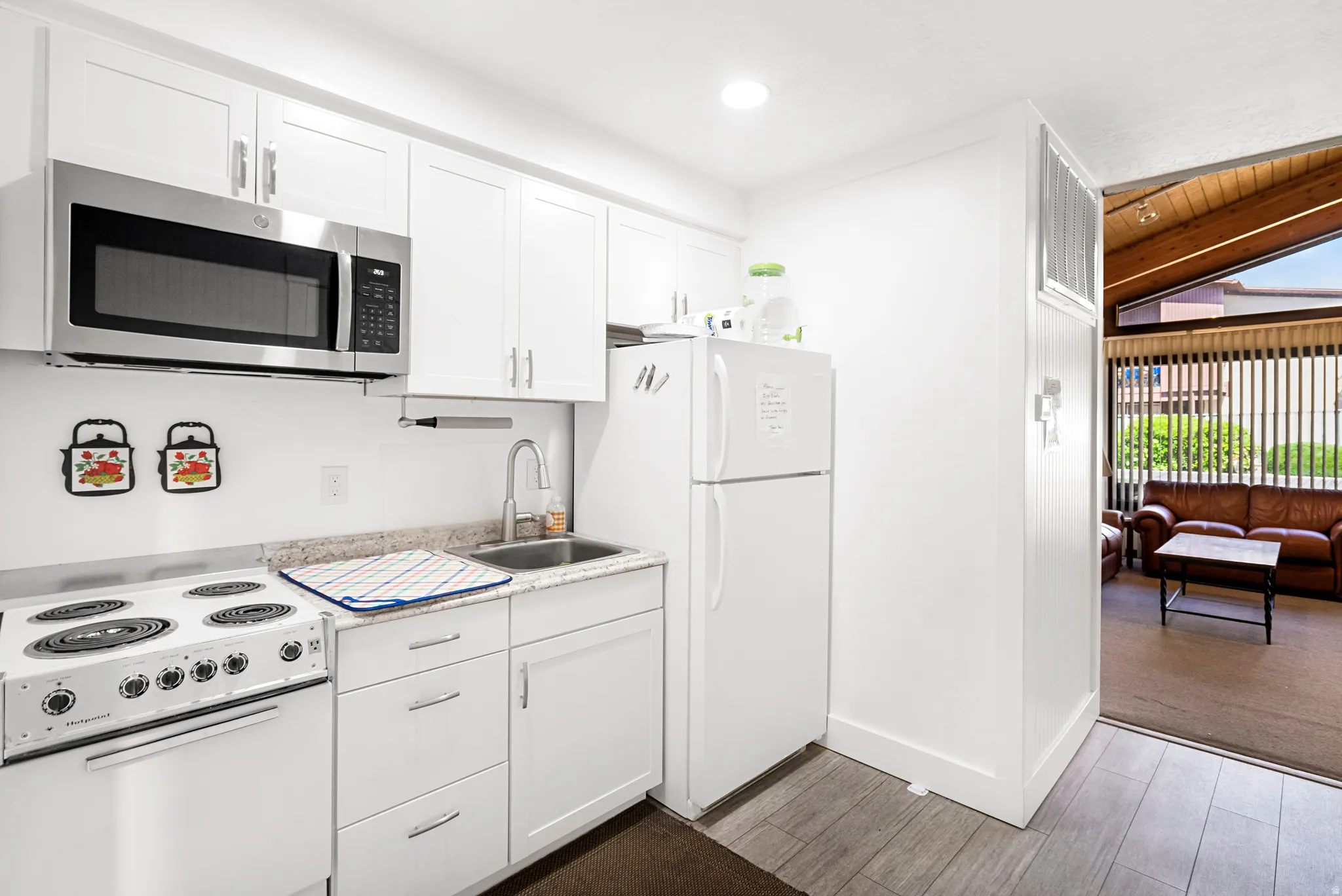 Compact kitchen featuring white cabinetry with brushed nickel hardware, a stainless steel over-the-range microwave, a white electric range, wood-finish flooring, and a white refrigerator