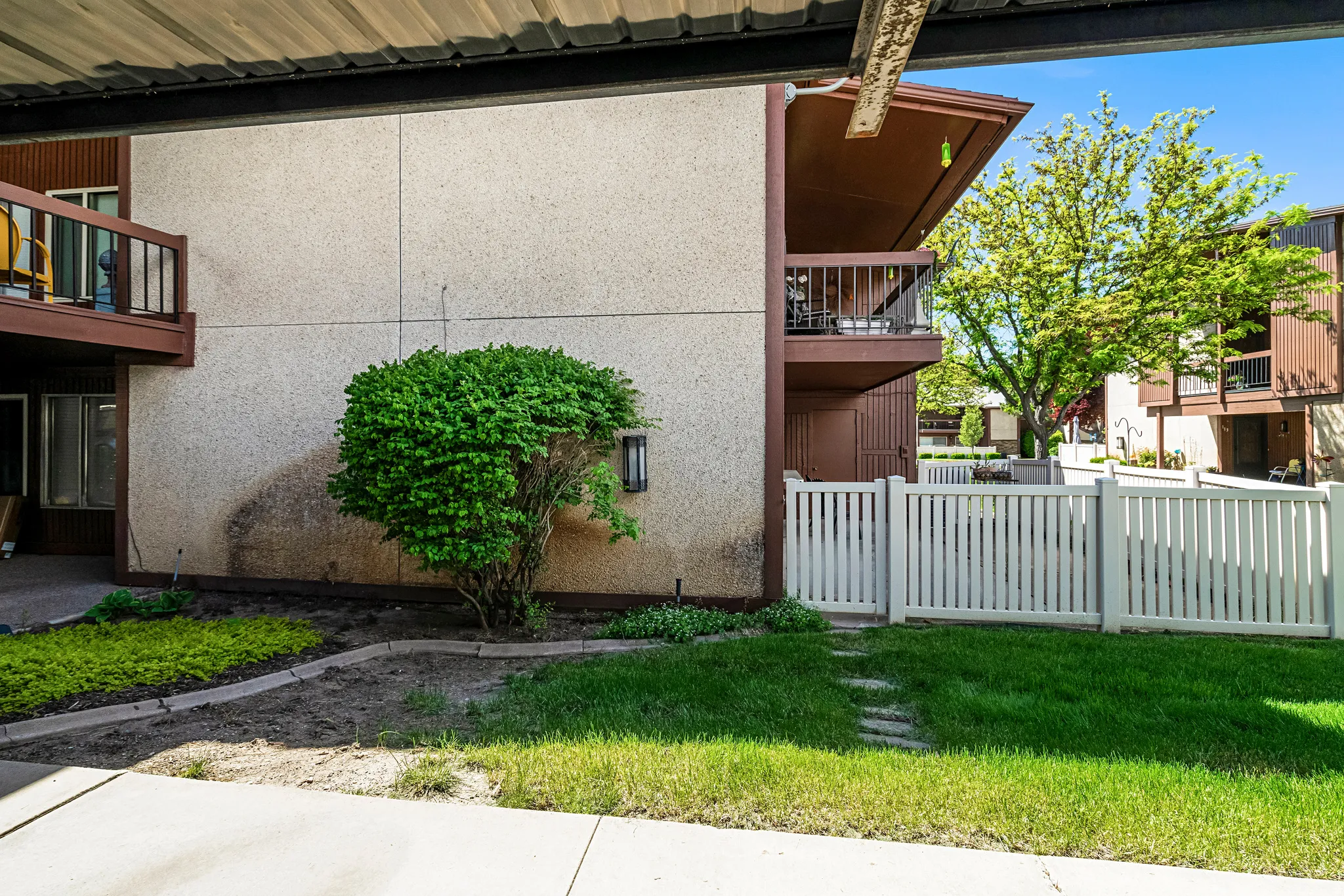 Private patio area with stucco exterior and white picket fencing, featuring a mature shrub, integrated wall lighting, and landscaping