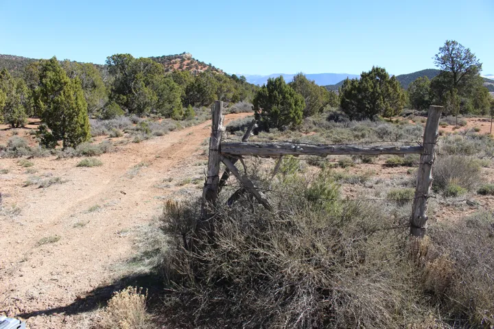 Expansive natural terrain featuring a dirt pathway, mature juniper trees, scattered brush, a rustic wood fence, and distant mountainous topography