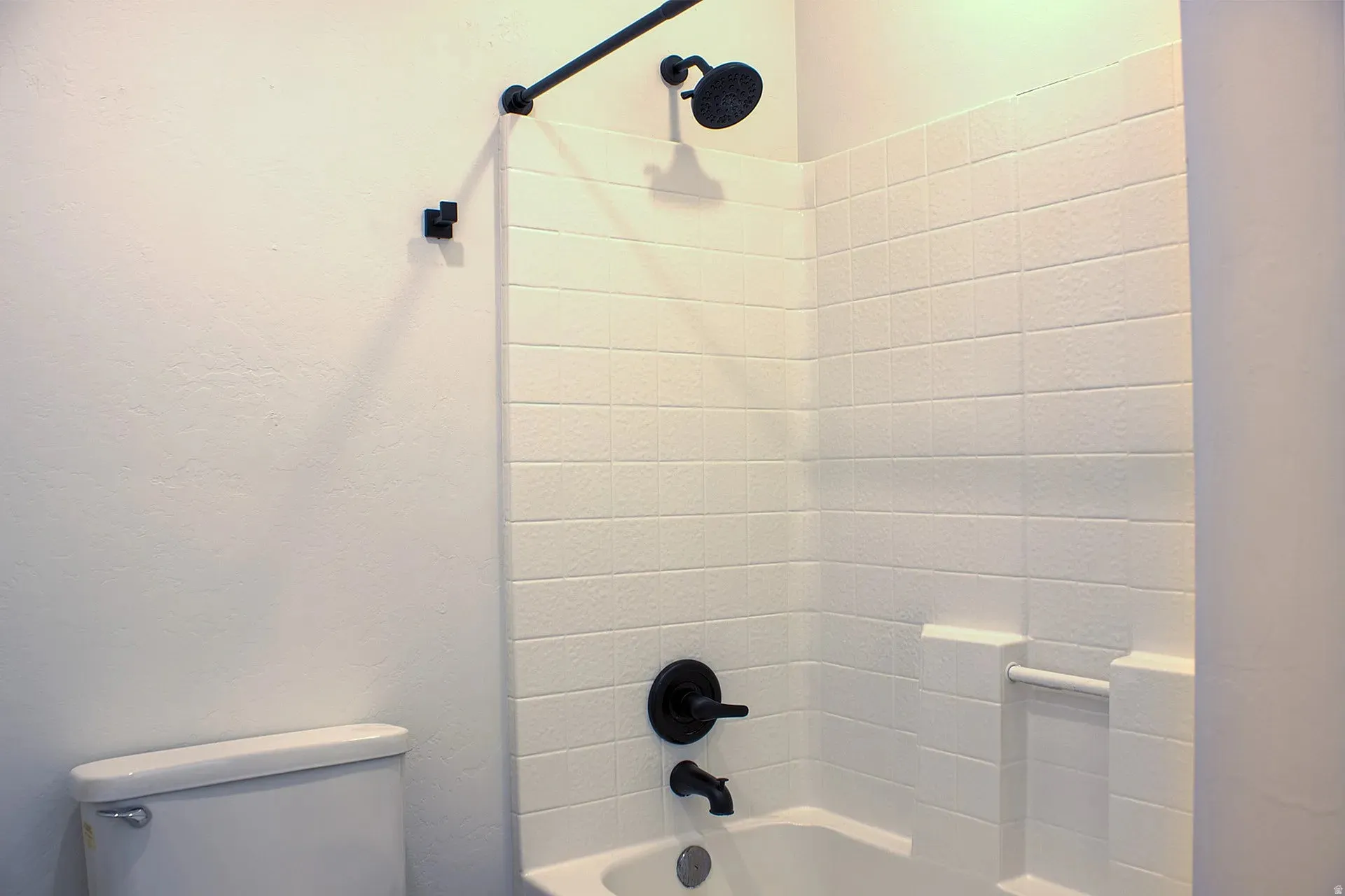 Bathtub and shower combination featuring white tiled walls, matte black shower fixtures, an integrated soap dish, and a white porcelain toilet
