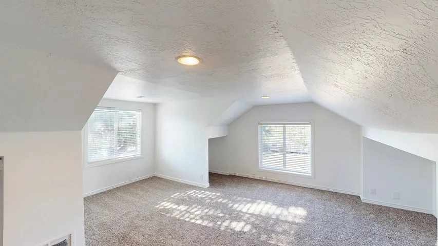 Spacious room featuring sloped ceilings, two windows with blinds, recessed lighting, and neutral-toned carpeting