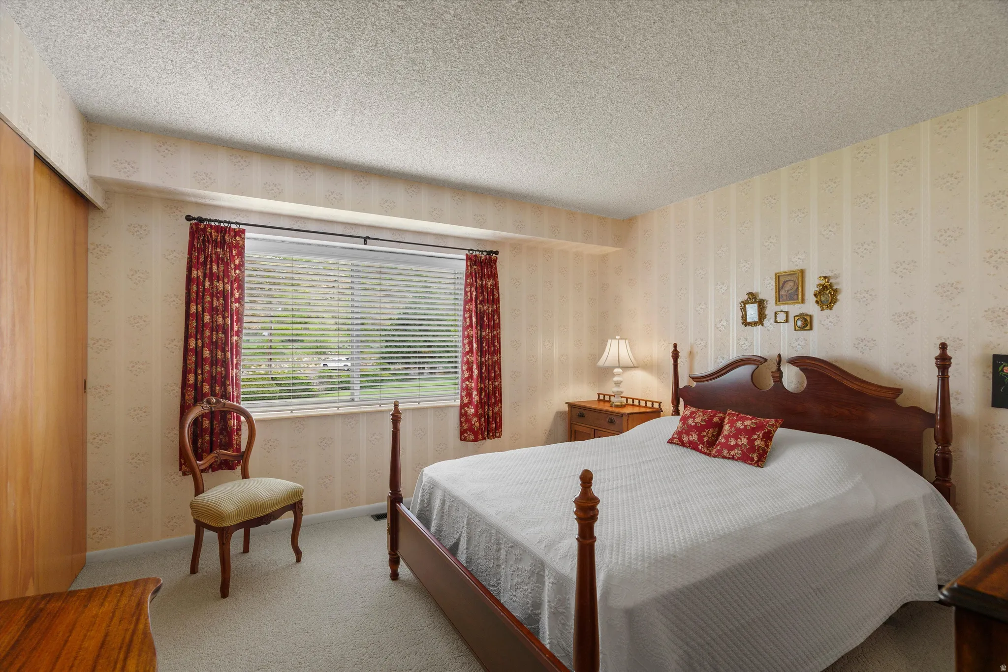 Room featuring a wide window with horizontal blinds, textured wall covering, light-colored carpeting, and a wood-finish bi-fold closet door
