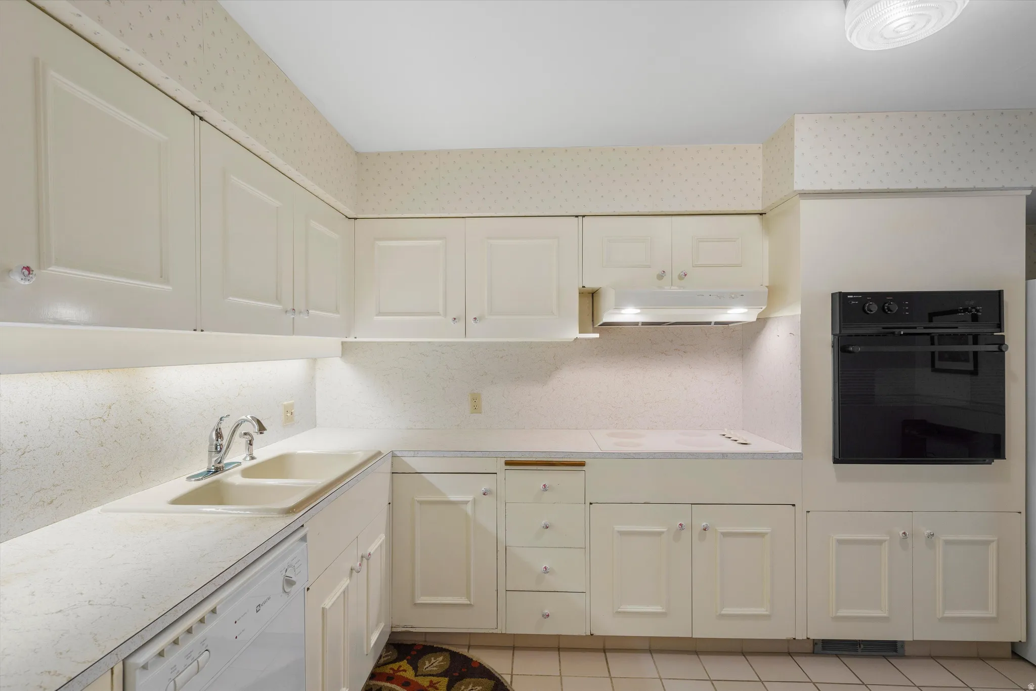 Kitchen featuring white cabinetry with panel detailing, light-toned countertops, a dual basin sink with chrome faucet, a built-in oven, and white tile flooring