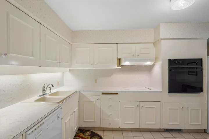 Kitchen featuring white cabinetry with panel detailing, light-toned countertops, a dual basin sink with chrome faucet, a built-in oven, and white tile flooring