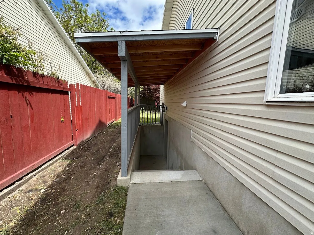 Covered concrete walkway leading to a basement entrance, featuring a wood-finish ceiling, metal railing, and vinyl siding exterior
