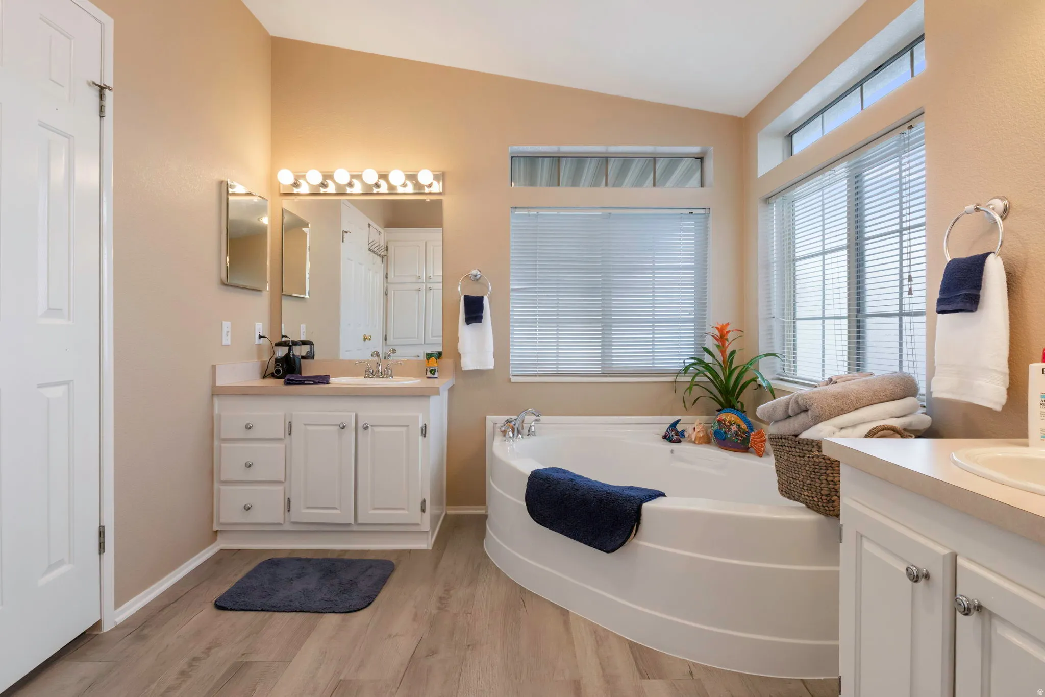 Bathroom featuring a corner soaking tub, dual vanity areas, wood-finish flooring, and multiple windows with blinds