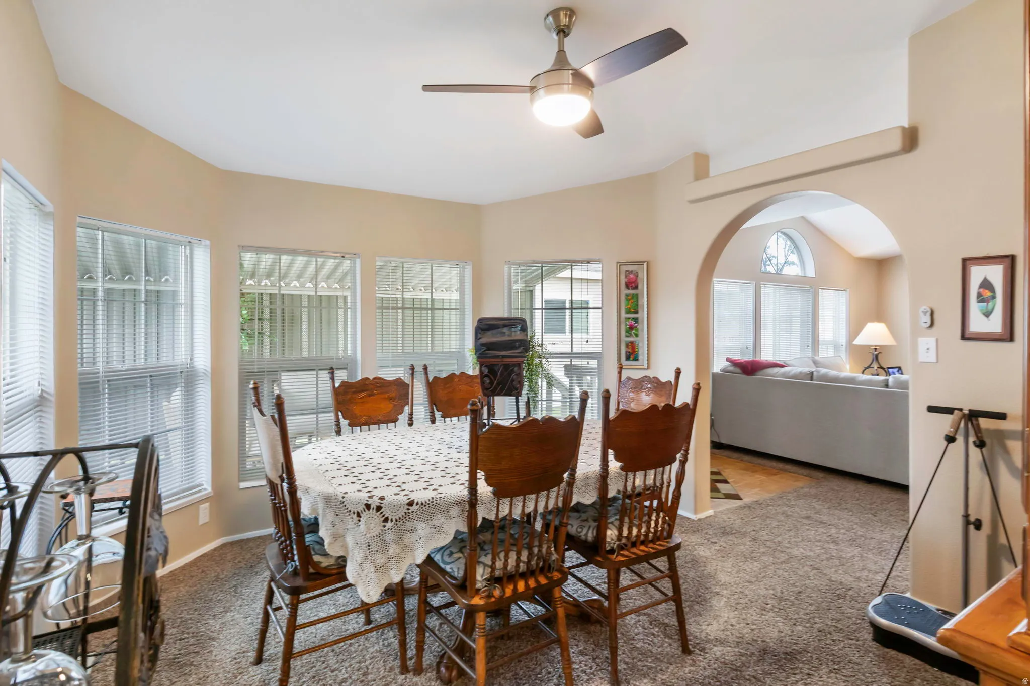 Carpeted dining area featuring multiple windows with blinds, a ceiling fan with integrated lighting, and a wide arched opening to an adjacent living space