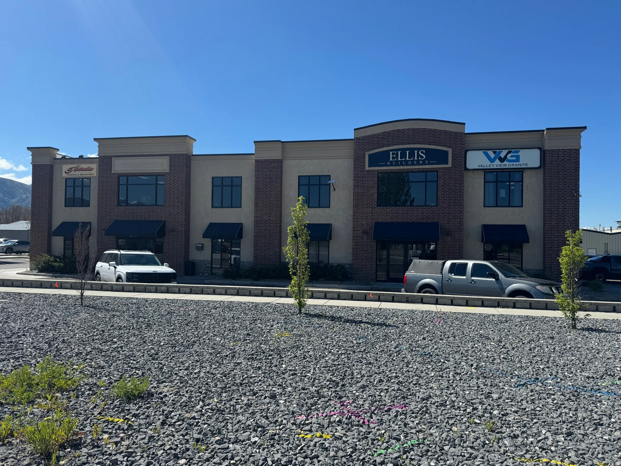 Two-story commercial building featuring brick and stucco facade, arched entryway, multiple window awnings, and an adjacent gravel parking area