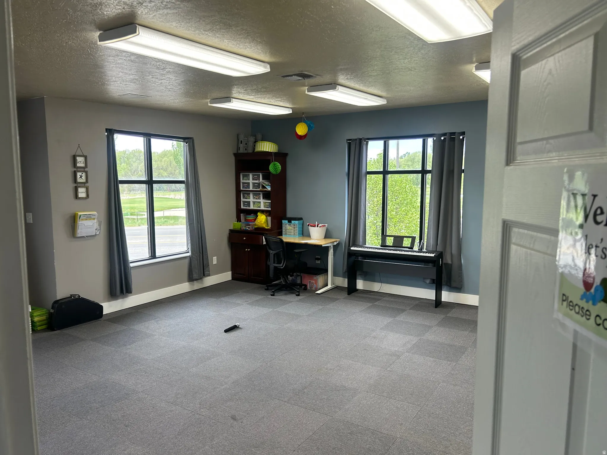 Interior space featuring checkerboard-patterned carpet tile flooring, two large windows with dark grey curtains, and two ceiling-mounted rectangular fluorescent light fixtures