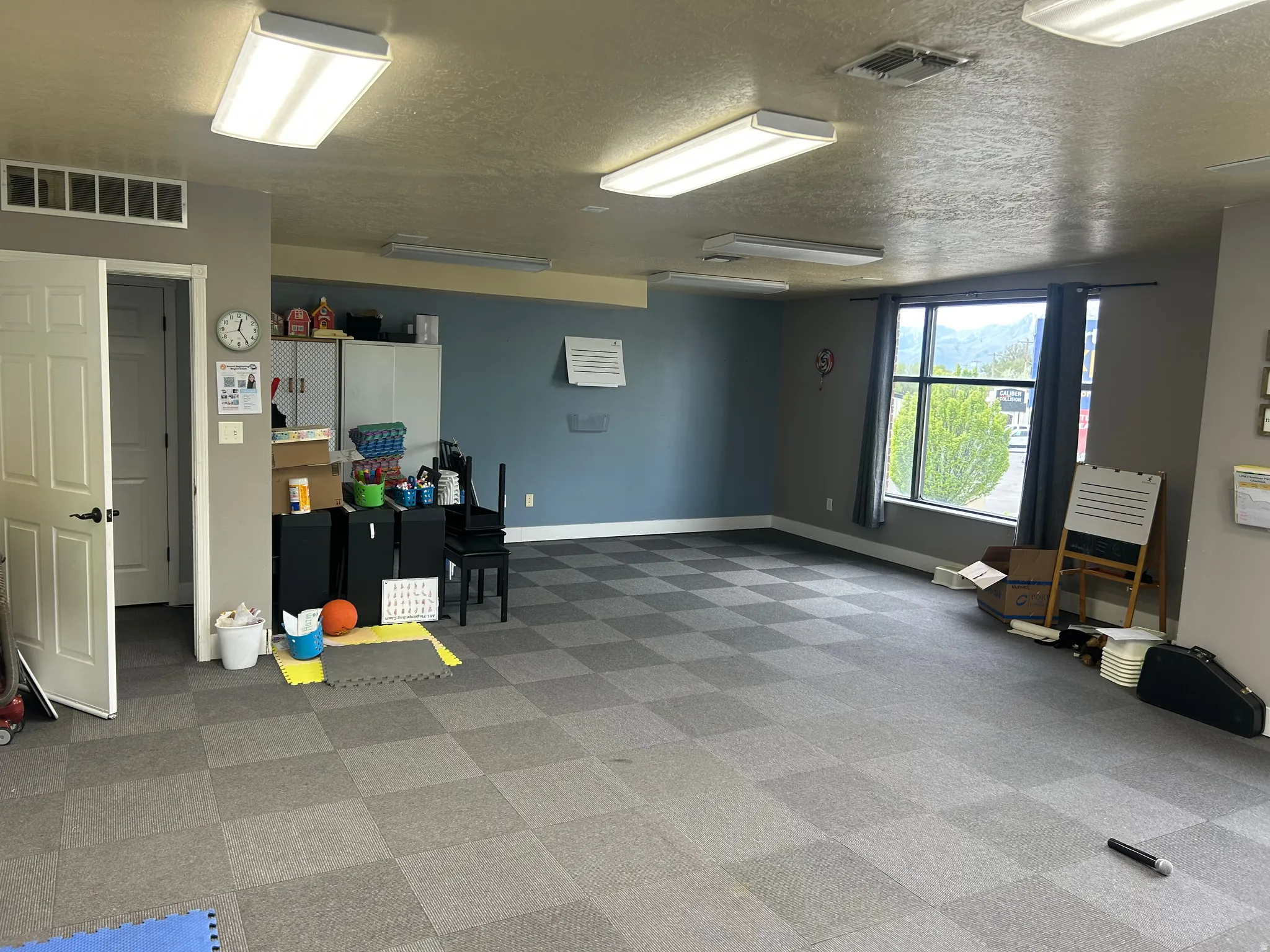 Spacious room featuring checkered carpet tile flooring, a large window, and multiple fluorescent ceiling lights
